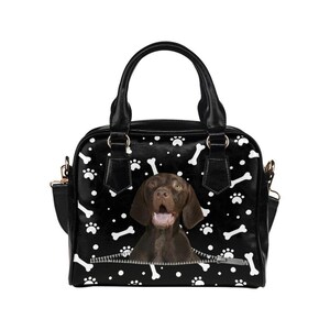May include: Black handbag with a dog design. The bag features a brown dog emerging from a zipper, set against a black background with white paw prints and bone illustrations. Includes a top handle and a shoulder strap.
