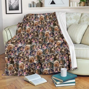 May include: A cozy blanket featuring a repeating pattern of brown and tan dogs with their tongues out. The blanket is draped over a sofa, with a white edge visible. The blanket is made of soft material and is perfect for snuggling.