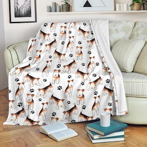 May include: White throw blanket featuring a repeating pattern of brown and white dogs and black paw prints. The blanket is draped over a sofa, with a portion folded over. The blanket is made of a soft material.