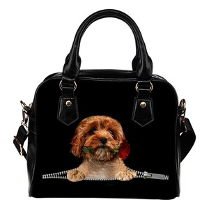 Könnte beinhalten: Schwarze Handtasche mit einem braunen und beigen Hund, der eine rote Rose hält. Der Hund scheint aus einem Reißverschluss zu kommen. Die Tasche hat einen oberen Griff und einen abnehmbaren Schultergurt.