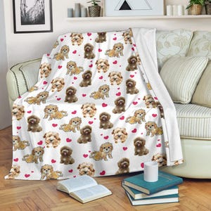 May include: White blanket featuring a repeating pattern of cartoon dogs in shades of brown and red hearts. The blanket is draped over a sofa, with a book and a mug visible in the foreground.