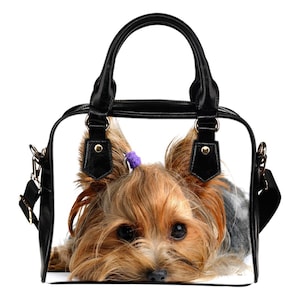 May include: Black and white handbag featuring a close-up of a Yorkshire Terrier dog. The dog has brown and tan fur, with a purple hair tie. The bag has black handles and a shoulder strap.