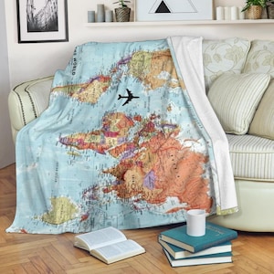 Può includere: Una coperta azzurra con un disegno della mappa del mondo e una grafica di un aereo nero. La mappa mostra i continenti in arancione, verde e marrone. La coperta è drappeggiata su un divano con cuscini bianchi e beige.