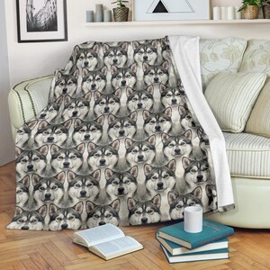 May include: A cozy throw blanket featuring a repeating pattern of Siberian Husky dog faces in shades of gray, black, and white. The blanket is draped over a sofa, showcasing the detailed watercolor-style illustrations of the dogs.