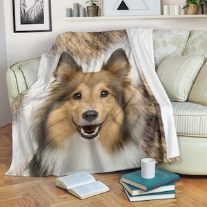 Puede incluir: Una manta blanca con una imagen de primer plano de un perro Sheltie sonriente. El perro tiene pelaje marrón y blanco, con la boca abierta. La manta está sobre un sofá con cojines estampados.