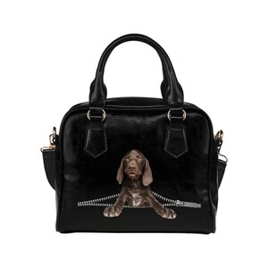 May include: Black handbag featuring a brown dog graphic peeking out from a zipper design. The bag has a structured shape with a top handle and a detachable shoulder strap. The dog has brown fur and is positioned as if emerging from the zipper.