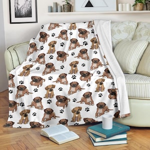 Peut inclure: Plaid blanc avec un motif répété d'illustrations de carlins bruns et d'empreintes de pattes noires. La couverture est drapée sur un canapé, mettant en valeur le design et la texture du tissu doux et confortable.