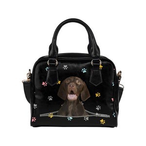 Puede incluir: Bolso de mano negro con un perro marrón saliendo de una cremallera. El bolso está adornado con huellas de patas de colores. Tiene un asa superior y una correa para el hombro desmontable.