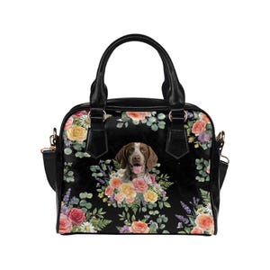 May include: Black handbag featuring a German Shorthaired Pointer surrounded by a floral arrangement of roses, daisies, and lavender. The bag has black handles and a detachable shoulder strap.