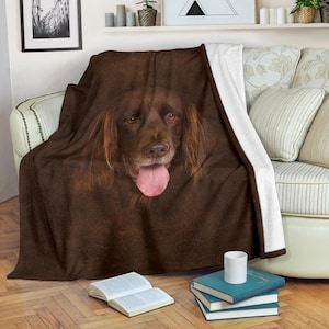 May include: A brown throw blanket featuring a close-up of a dog's face with its tongue out. The dog has brown fur and eyes. The blanket is draped over a couch with white and floral patterned pillows.