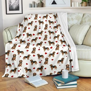 Puede incluir: Manta blanca con un patrón repetido de perros salchicha marrones y negros y corazones rojos. La manta está sobre un sofá, mostrando el diseño y la textura suave. Un artículo de decoración del hogar acogedor.