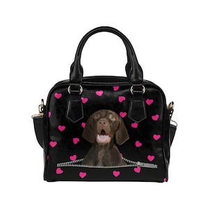 May include: Black handbag with pink hearts and a brown dog graphic. The dog appears to be emerging from a zipper. The bag has a top handle and a detachable shoulder strap.
