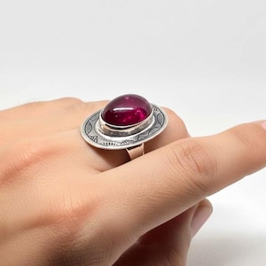 May include: A silver ring featuring a large, oval, deep red gemstone set in a circular, engraved setting. The ring has a unique, eye-catching design, with intricate patterns around the gemstone. The ring is shown on a hand.
