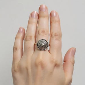 Può includere: Un anello argentato con un design circolare intrecciato. Il centro dell'anello presenta un motivo dettagliato e strutturato, circondato da un bordo a forma di corda ritorta. L'anello è indossato su un dito, mostrando la sua intricata fattura.