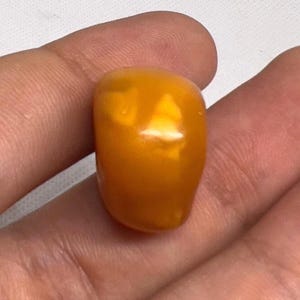 Puede incluir: Una gema pulida de color ámbar, con forma suave y redondeada. La piedra tiene un tono dorado cálido y una superficie brillante, que refleja la luz. La gema se sostiene en una mano, mostrando su tamaño y color.