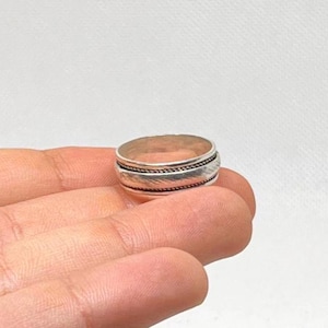 May include: A silver ring with a textured design. The ring features a band of braided detail between two smooth bands. The ring is held in a hand against a white background.