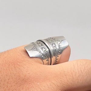 May include: A silver ring with intricate geometric patterns. The ring has a wide band and a shield-like design on the top, featuring detailed lines and shapes. The ring is worn on a finger.