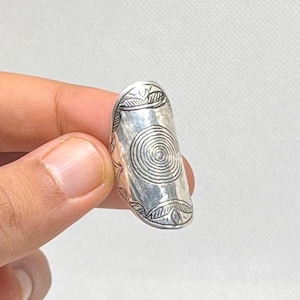 Könnte beinhalten: Ein silberfarbener Ring mit länglichem Design. Der Ring hat ein eingraviertes Spiralmuster in der Mitte und blattartige Designs an den Rändern. Der Ring wird von den Fingern einer Person vor einem weißen Hintergrund gehalten.