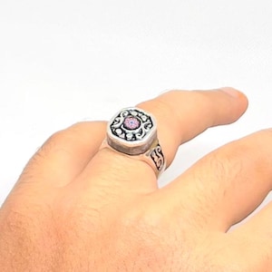 Può includere: Un anello d'argento con una faccia quadrata ornata con una pietra rosa centrale circondata da un motivo decorativo. L'anello è indossato su un dito, mostrando il suo design e la sua maestria intricati. La fascia ha un design a volute dettagliato.