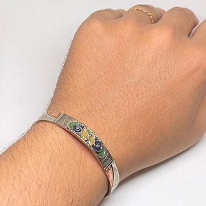 May include: A silver and copper bracelet with a green, yellow and blue enamel design. The bracelet is on a person's wrist.