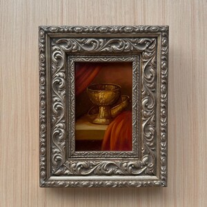 May include: A rectangular painting in a silver-toned ornate frame with scrollwork details. The artwork depicts a golden chalice resting on a flat surface, positioned next to draped red fabric that creates deep shadows and highlights in the scene.
