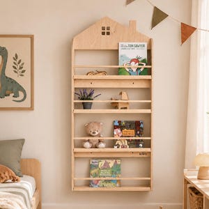 Könnte beinhalten: Ein helles, wandmontiertes Bücherregal aus Holz in Hausform mit Schornstein und einem kleinen quadratischen Fensterausschnitt. Das Regal verfügt über vier horizontale Ebenen mit verschiedenen Kinderbüchern, einer kleinen Topfpflanze und einem Teddybären.
