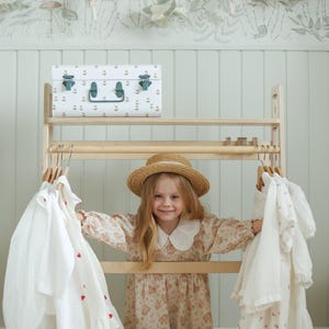 Pode incluir: Um cabideiro de madeira clara com duas barras horizontais para pendurar roupas. A prateleira superior cont&eacute;m um ba&uacute; de armazenamento de metal branco com um padr&atilde;o floral e fechos verdes. V&aacute;rias camisas e vestidos brancos est&atilde;o pendurados.
