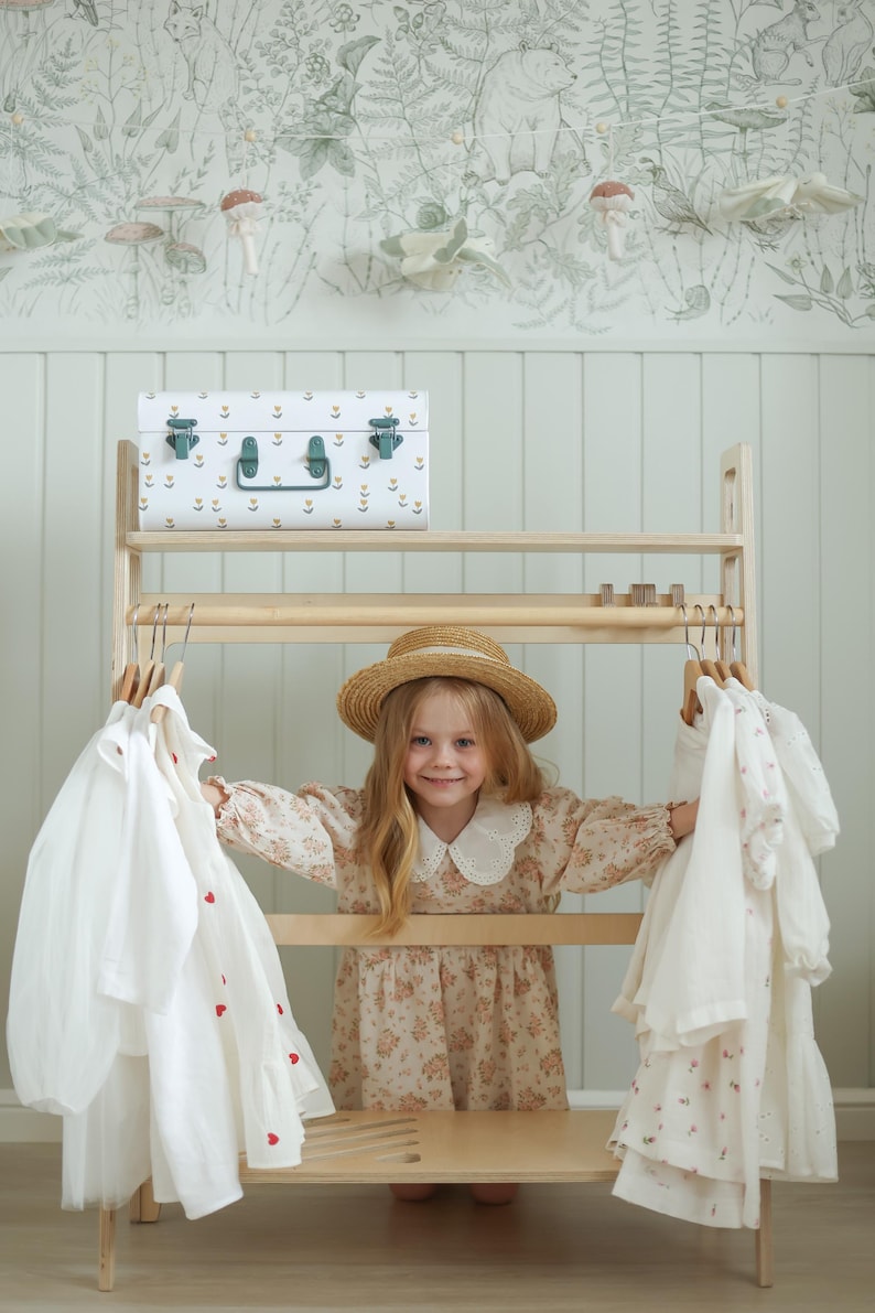 Pode incluir: Um cabideiro de madeira clara com duas barras horizontais para pendurar roupas. A prateleira superior cont&eacute;m um ba&uacute; de armazenamento de metal branco com um padr&atilde;o floral e fechos verdes. V&aacute;rias camisas e vestidos brancos est&atilde;o pendurados.