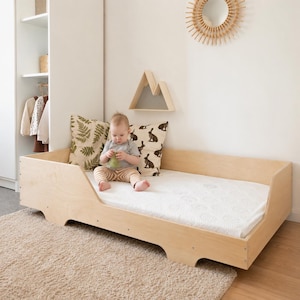 Pode incluir: Uma estrutura de cama para criança em madeira clara de perfil baixo com colchão branco, apresentando um design lateral recortado. A cama é decorada com uma almofada com padrão de fetos verdes e uma almofada creme com ilustrações de coelhos.