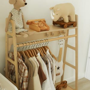 Pode incluir: Um cabideiro de madeira clara com uma prateleira superior contendo um boneco de cachorro de pel&uacute;cia, um telefone de brinquedo de madeira e um rinoceronte de pel&uacute;cia sobre rodas. Abaixo, uma haste sustenta camisas e su&eacute;teres em cabides.