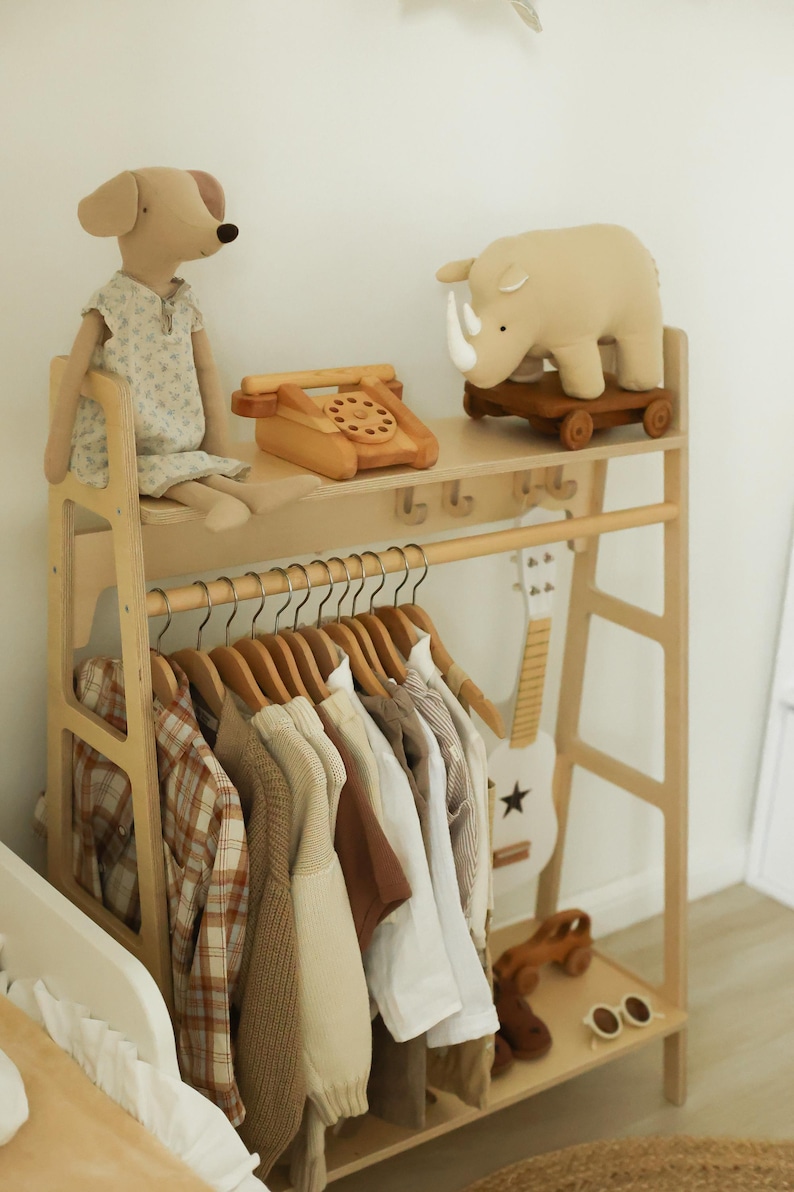 Pode incluir: Um cabideiro de madeira clara com uma prateleira superior contendo um boneco de cachorro de pel&uacute;cia, um telefone de brinquedo de madeira e um rinoceronte de pel&uacute;cia sobre rodas. Abaixo, uma haste sustenta camisas e su&eacute;teres em cabides.