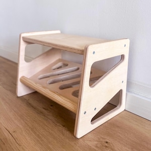 Puede incluir: Un taburete de dos niveles de madera natural. La parte superior es un rectángulo sólido, mientras que la parte inferior tiene formas recortadas. Los lados presentan cortes angulados, y el taburete está ensamblado con tornillos. Diseñado para niños.
