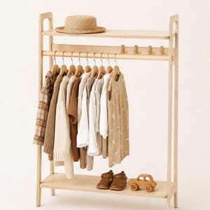 Op de afbeelding: Een kledingrek van lichtgekleurd hout met een bovenplank met een strooien hoed, een roede met diverse kledingstukken op houten hangers, een rij kleine haakjes en een onderste plank met een paar bruine leren schoenen en een houten speelgoedauto.