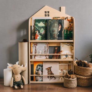 Könnte beinhalten: Ein helles, hausförmiges Bücherregal aus Holz mit drei Ebenen, in denen verschiedene illustrierte Kinderbücher stehen. Die Regale verfügen über horizontale Schienen, um die Bücher aufrecht zu halten. Ein Plüsch-Hirsch und geflochtene Körbe stehen daneben.