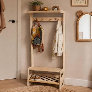 Pode incluir: Um cabideiro de madeira clara com prateleira superior, barra horizontal com ganchos e banco inferior com ripas para sapatos. O cabideiro sustenta uma mochila bege com estampa de dinossauro, uma camisa creme e tênis marrons.