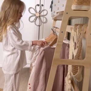 Pode incluir: Um cabideiro de madeira clara com v&aacute;rias prateleiras e ganchos. O cabideiro sustenta v&aacute;rias pe&ccedil;as de roupa em cabides de madeira, incluindo um vestido liso em rosa seco e um vestido branco com padr&otilde;es de cora&ccedil;&otilde;es vermelhos, al&eacute;m de um cesto.