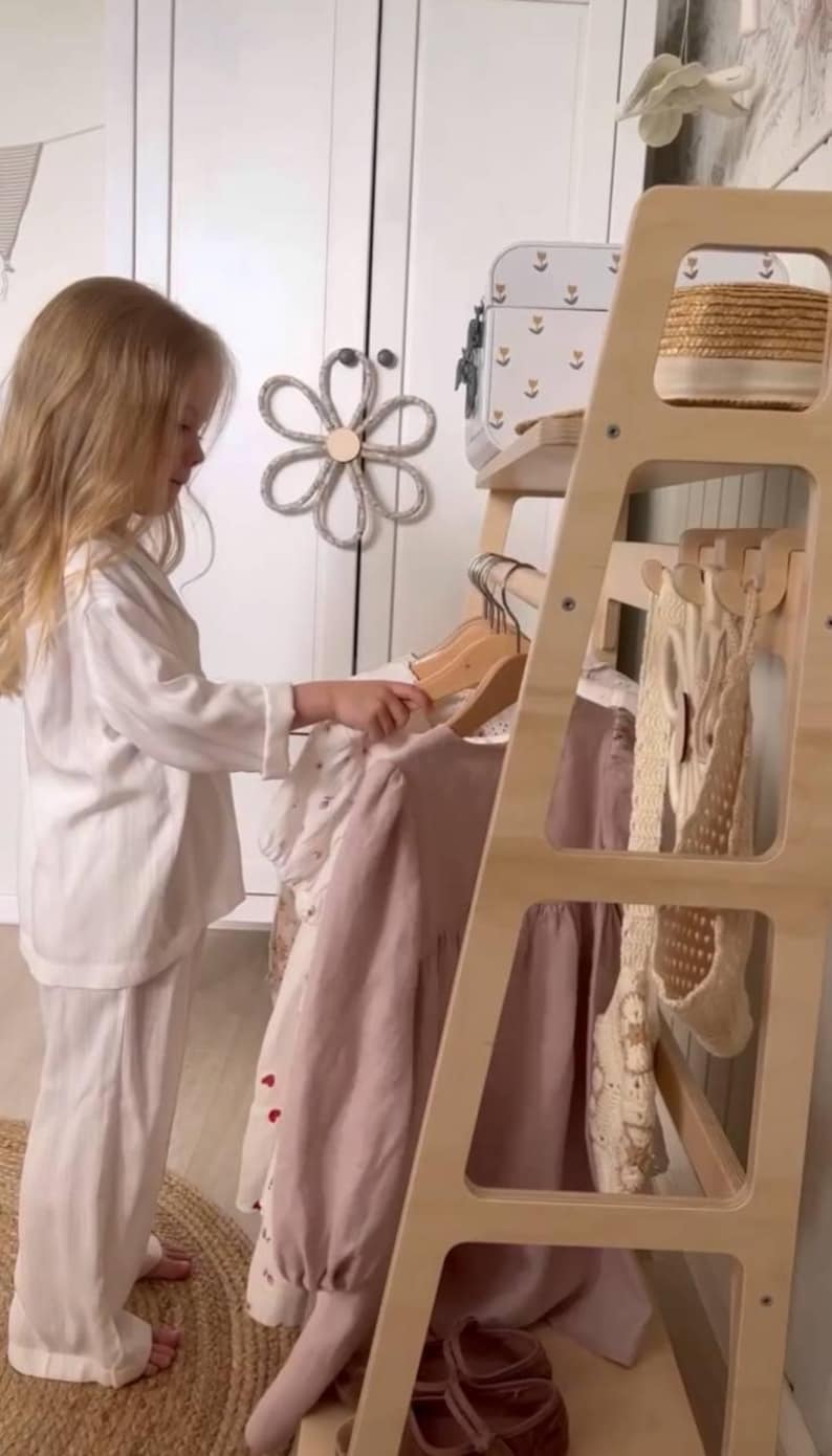 Pode incluir: Um cabideiro de madeira clara com v&aacute;rias prateleiras e ganchos. O cabideiro sustenta v&aacute;rias pe&ccedil;as de roupa em cabides de madeira, incluindo um vestido liso em rosa seco e um vestido branco com padr&otilde;es de cora&ccedil;&otilde;es vermelhos, al&eacute;m de um cesto.