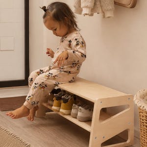 May include: A light-coloured wooden shoe bench with a flat top and a lower shelf. The bench features side panels with triangular cutouts and is constructed with visible metal screws. It is designed for storage and seating in an entryway space.