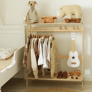 Pode incluir: Um cabideiro de madeira clara com uma prateleira superior contendo um c&atilde;o de pel&uacute;cia, um telefone de brinquedo de madeira e um rinoceronte de pel&uacute;cia sobre rodas. O cabideiro sustenta v&aacute;rias roupas infantis, uma guitarra branca e um carro de madeira.