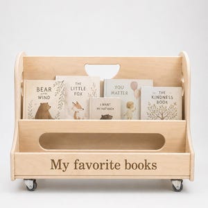 Könnte beinhalten: Ein helles Bücherregal aus Holz auf Rollen mit zwei Ablagen. Die Vorderseite ist mit dem eingravierten Text "My favorite books" versehen. In den Regalen stehen mehrere Kinderbücher mit den Titeln "Bear and the Wind", "The Little Fox", "I Want My Hat Back", "You Matter" und "The Kindness Book".