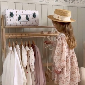 Pode incluir: Um cabideiro de madeira sustenta v&aacute;rias pe&ccedil;as em cabides, incluindo roupas brancas e malvas de manga comprida. Um ba&uacute; de armazenamento branco com padr&otilde;es repousa na prateleira superior. Uma pessoa veste um vestido floral e chap&eacute;u de palha.