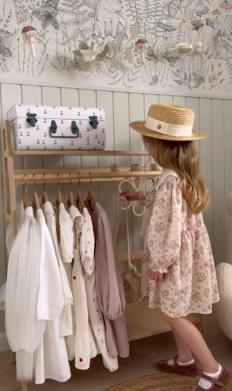 Pode incluir: Um cabideiro de madeira sustenta v&aacute;rias pe&ccedil;as em cabides, incluindo roupas brancas e malvas de manga comprida. Um ba&uacute; de armazenamento branco com padr&otilde;es repousa na prateleira superior. Uma pessoa veste um vestido floral e chap&eacute;u de palha.