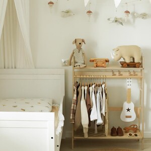 Pode incluir: Uma cama infantil branca com dossel, um cabideiro de madeira com v&aacute;rias pe&ccedil;as de roupa, um peluche de c&atilde;o, um telefone de brinquedo de madeira, um rinoceronte sobre rodas, uma guitarra de brinquedo branca, sapatos castanhos, um carro de madeira e &oacute;culos.