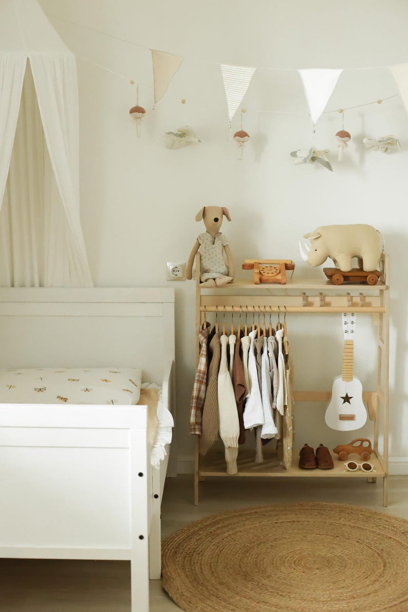 Pode incluir: Uma cama infantil branca com dossel, um cabideiro de madeira com v&aacute;rias pe&ccedil;as de roupa, um peluche de c&atilde;o, um telefone de brinquedo de madeira, um rinoceronte sobre rodas, uma guitarra de brinquedo branca, sapatos castanhos, um carro de madeira e &oacute;culos.