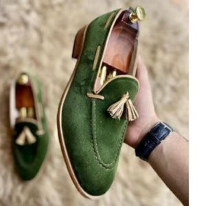 May include: Green suede loafers with a tassel detail. The shoes have a rounded toe and a low heel. The image also shows a second shoe in the background. The shoes are on a fluffy, light-colored surface.