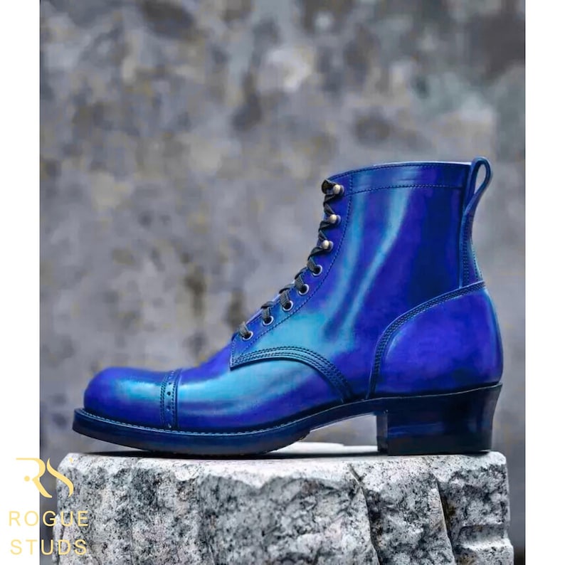 May include: A blue leather boot with a cap toe and lace-up closure. The boot has a stacked heel and a pull tab at the back. The boot is displayed on a stone surface. The brand name "ROGUE STUDS" is visible.