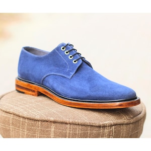 May include: A single blue suede shoe with black laces and gold eyelets. The shoe has a brown sole with a black trim and is displayed on a brown patterned cushion. The shoe is a classic design.