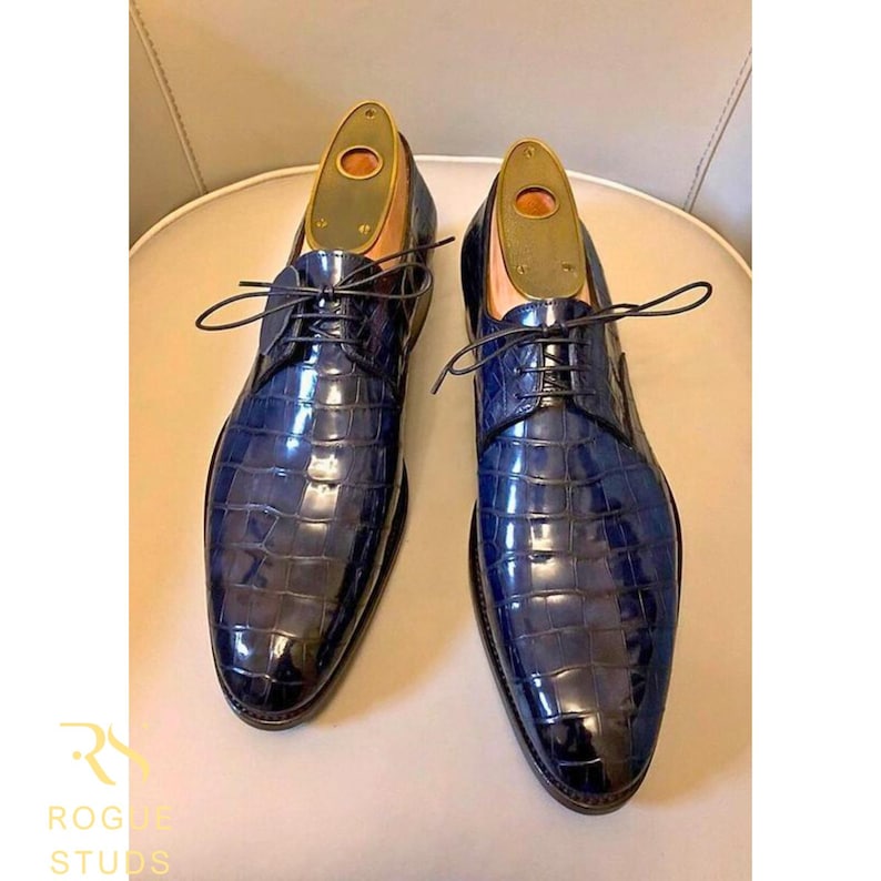 May include: A pair of navy blue leather dress shoes with a crocodile skin pattern. The shoes have black laces and are displayed with wooden shoe trees inside. The shoes are on a light-colored surface.