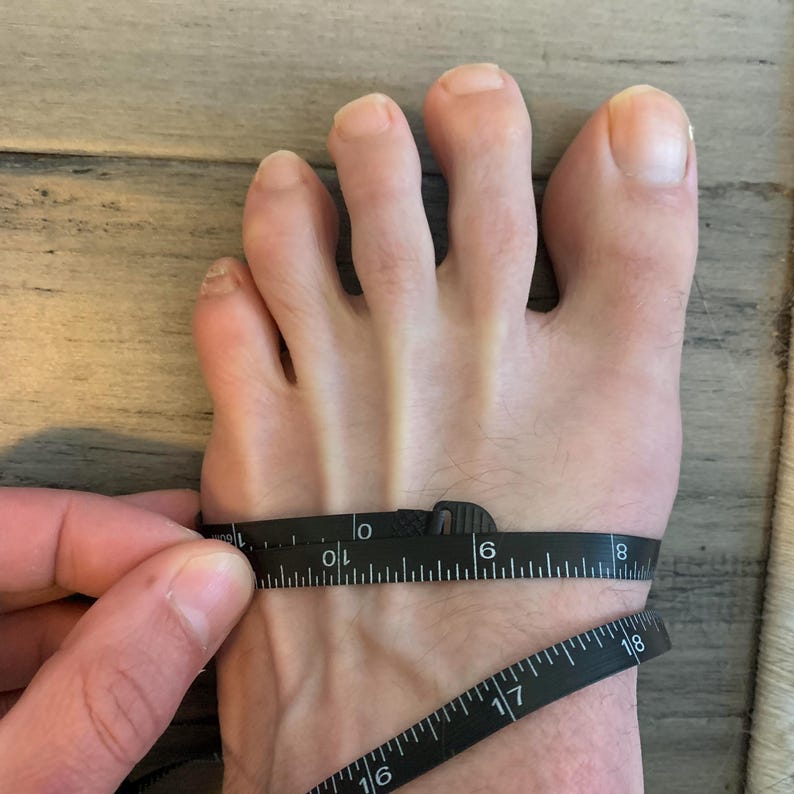 May include: A foot is being measured with a black measuring tape. The tape is wrapped around the foot, showing the width. The foot is pale with visible toes and toenails. The background is a light wood grain.