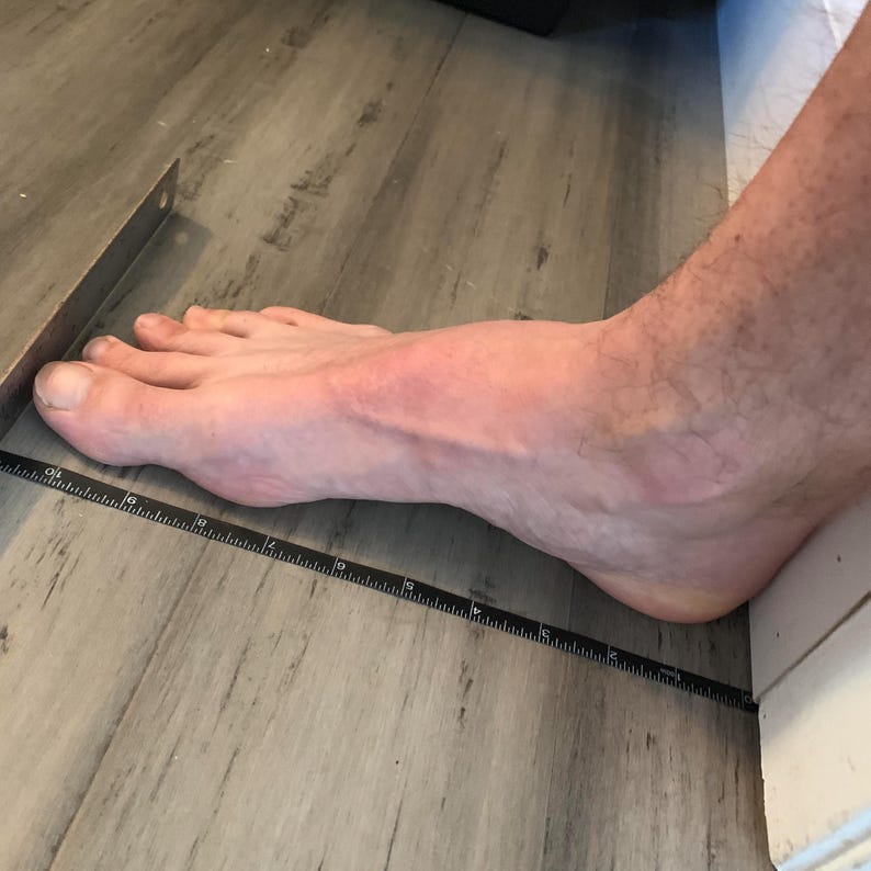May include: A foot is placed next to a ruler to measure its length. The foot is positioned against a wall, and the ruler extends along the foot's side. The floor is a light gray wood-like pattern.